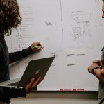 A woman holding a laptop writing on a whiteboard and a man observing the lecture.